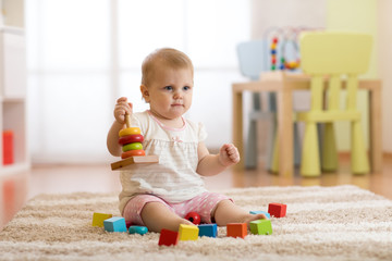 Cute baby girl playing with colorful toys sitting on carpet in white sunny bedroom. Child with educational toys. Early development.