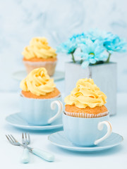 Cupcake with tender yellow cream decoration and bouquet of blue chrysanthemum in retro shabby chic vase on blue pastel background.