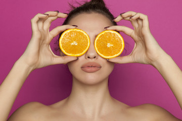 Young sexy woman posing with slices of oranges on her face on pink background