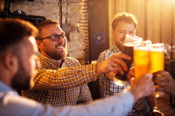 Young satisfied bearded men celebrating and toasting with a beer in the sunny pub after work.