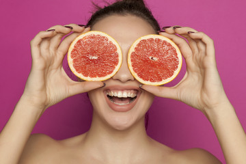 Happy young woman posing with slices of red grapefruit on her face on pink background