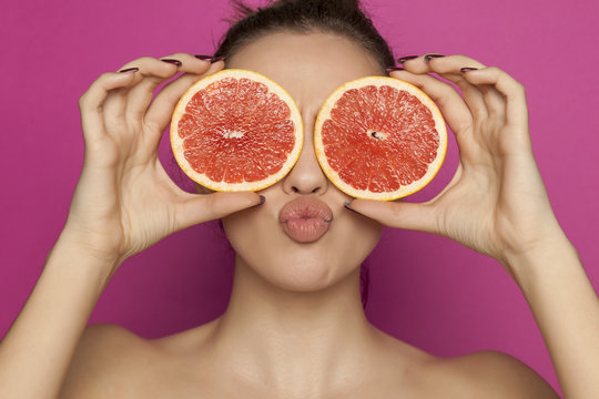 Young sexy woman posing with slices of red grapefruit on her face on pink background