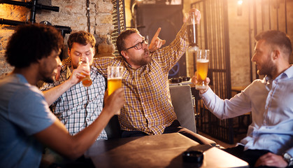 Multicultural cheerful drunk friends sitting hugged in the bar and celebrating with a beer.