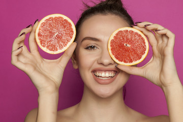 Happy young woman posing with slices of red grapefruit on her face on pink background