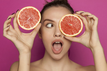 Young surprised woman posing with slices of red grapefruit on her face on pink background
