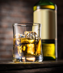 Glass of whiskey with ice cubes and whiskey bottle on wooden table.