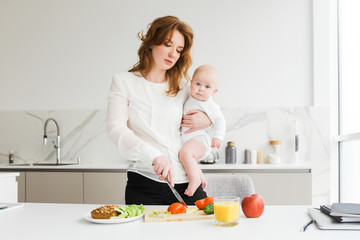 Portrait of beautiful mother holding her cute little baby while standing and cooking on kitchen isolated
