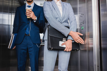 Close-up view of businesswoman pushing button in elevator by man in earphones with coffee cup