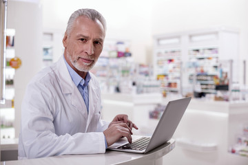 Pharmacy organization. Mature earnest male pharmacist working on laptop while looking at camera