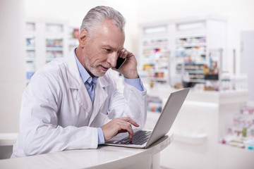 Important call. Senior handsome male pharmacist male pharmacist working on laptop while making call