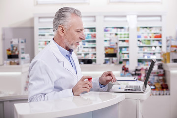 Naklejka na ściany i meble Let me check. Senior attentive male pharmacist posing in profile while typing on laptop
