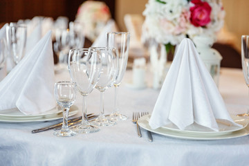 Glasses, flowers, fork, knife, napkin folded in a pyramid, served for dinner in restaurant with cozy interior. Wedding decorations and items for food, arranged by the catering service on a large table