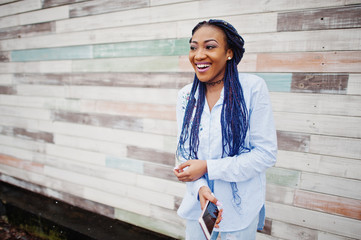 Stylish african american girl with dreads holding mobile phone at hand, outdoor against wooden wall.