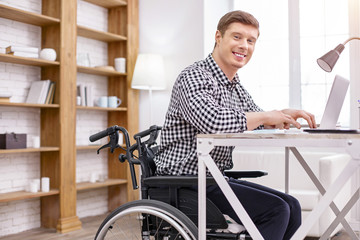 Join me. Delighted differently abled man sitting on the wheelchair and working with computer