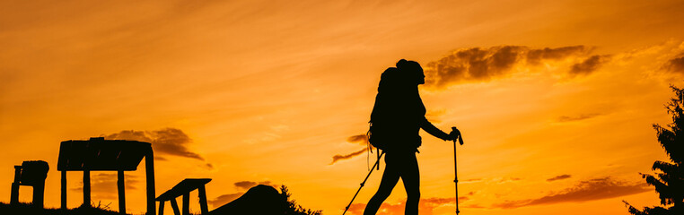 Silhouette traveler woman hiking with backpack and trekking pole, sunset orange sky on the background