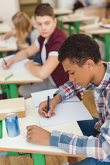 Elevated view of african american teenage schoolboy writing in textbook and classmates on behind