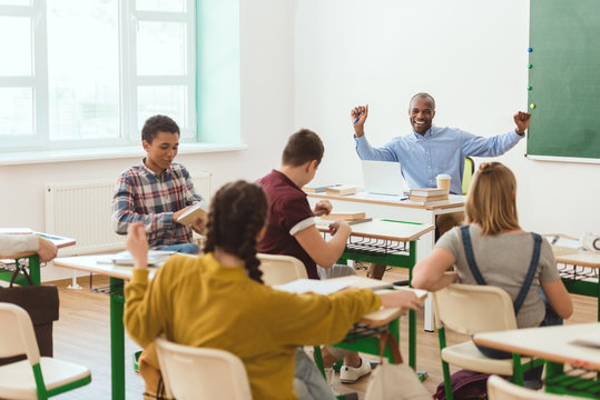 Multicultural group of teenage schoolchildren and smiling african american teacher with raising arms