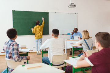 Rear view of schoolgirl writing on chalk board and classmates with teacher sitting in classroom