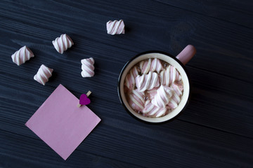 The pink mug of hot coffee with marshmallow and empty pink paper on a dark blue wooden desk.