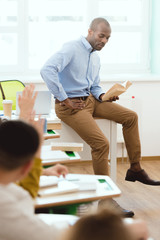 African american teacher sitting on table and reading book to schoolchildren with arms up in classroom