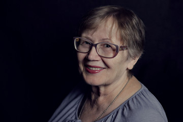 portrait of elderly woman with short blonde hair, wearing glasses and blue jacket on black background