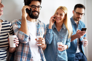 Close up view of young joyful people using phones while having a break and drinking coffee from a paper cups and laughing.