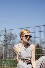 Natural looking woman with blonde short hair and tattoo, wearing sunglasses and grey training suit, sitting in sunny day in roller sport park