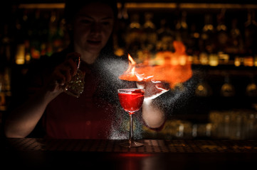 Female bartender spraying on the fire match above the cocktail in the dark