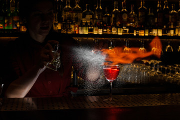 Female bartender spraying on the fire match above the cocktail on the dark blurred background