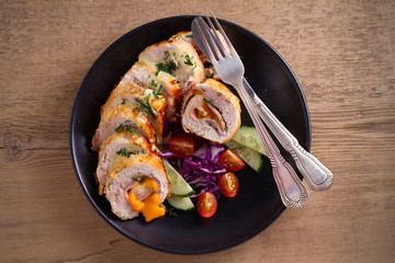 Chicken wrapped with ham and cheddar cheese; vegetables in black plate on wooden table. horizontal