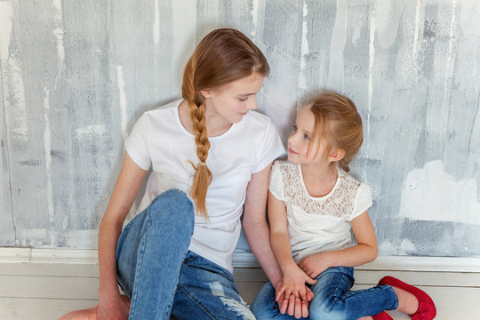 Two Happy Kids Sitting Against Grey Textured Wall Background And Embracing. Adorable Pretty Little Girl Hugging Tight Cute Teenage Girl, Showing Her Love And Care. Sisters Having Fun At Home