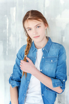 Young happy cute teenage girl in jeans, denim jacket and white T-shirt sitting on chair against grey textured wall background. Casual daily lifestyle. Schoolgirl student education and school concept