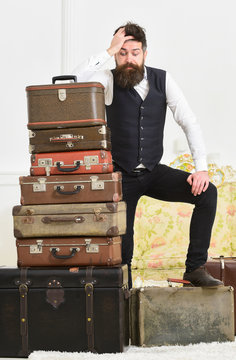 Macho elegant on disappointed face standing near pile of vintage suitcase. Luggage and travelling concept. Man, butler with beard and mustache delivers luggage, luxury white interior background.
