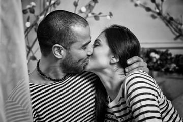 Black and white photo of charming international couple in striped sweaters kissing and hugging while sitting on the bed