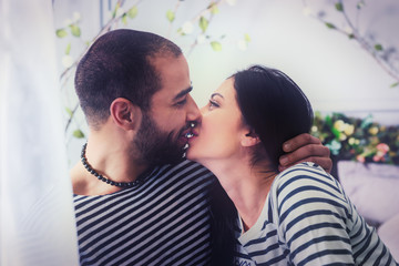 Portrait of adorable international couple in striped sweaters kissing and hugging while sitting on the bed