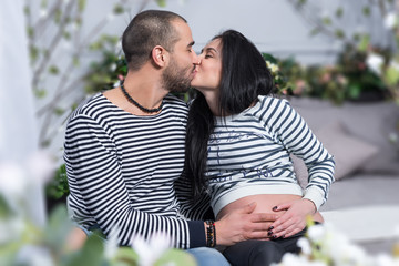 Obraz premium Happy international couple in striped sweaters is kissing, muslim man hugging belly of his pregnant wife while sitting on the bed