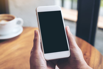 Mockup image of hands holding white mobile phone with blank black screen and coffee cup on wooden table in cafe