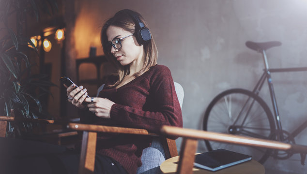 Young attractive girl wearing eye glasses and casual clothes listening digital music in headphones on smartphone at modern place.