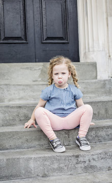 Emotional portrait of a sad, unhappy little girl with blond curly hair sitting on the front steps of her home in an urban setting. Cute expression and adorable face. child behavior concept photo