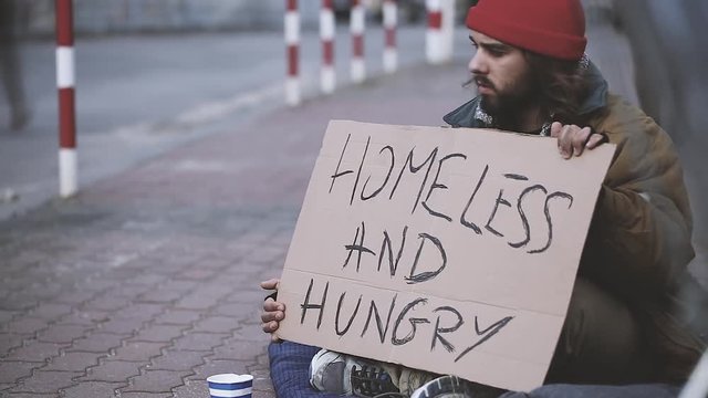 Time lapse video of a homeless and hungry beggar sitting on the sidewalk on which pedestrians are walking in the city