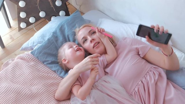 Young attractive blond mom take a photo in mobile phone with her little charming daughter in pink dresses. Family hug and kiss.
