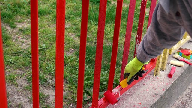 a worker with yellow gloves and paint roller painting a red fence on the background green grass