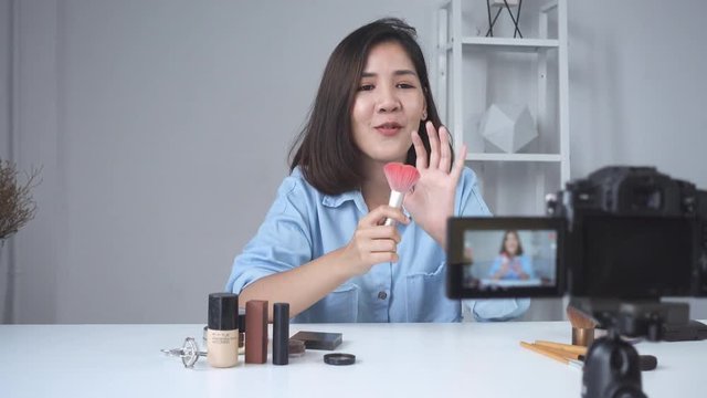 Happy smiling asian woman or beauty blogger with brush and camera recording video and waving hand at home. Beauty videoblog blogging people concept. Dolly shot.