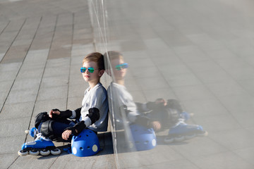 Young boy sitting near grey wall with inline roller skates and all protection at outdoor skate park