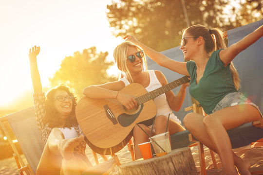 Group of young female friends sitting on beach on sunbeds,singing and playing guitar.Joying in sunset.