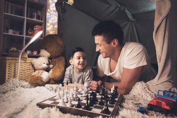 Father teaches little son how to play chess at night at home.