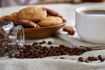 Coffee cup, jar with coffee beans, cookies over homespun tablecloth, selective focus, close-up, side view