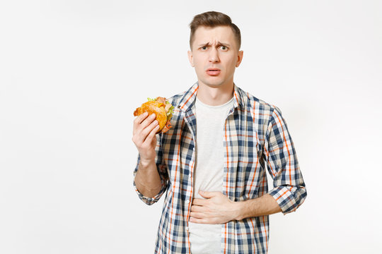 Illness sad young man in shirt put hand on pain abdomen, stomach-ache, standing and holding burger isolated on white background. Proper nutrition or American classic fast food. Area with copy space.