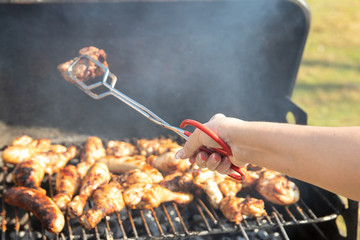 Woman is grilling steaks, grill sausages and chicken wings and using barbecue tongs. Closeup of hand outdoor. Concept food.