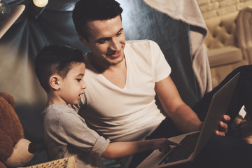 Father and son are watching video on laptop at night at home. - obrazy, fototapety, plakaty